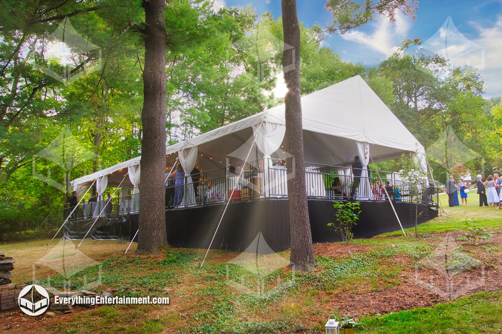 A 30x60 white wedding frame tent set up on an elevated leveled platform with safety railings and black skirting on a sloped backyard.