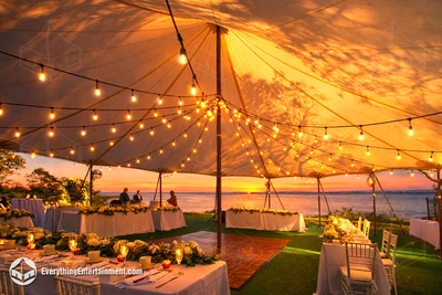 Sunset outdoor wedding reception under a glowing sailcloth tent with illuminated string lights, elegant white floral table settings, wooden dance floor, and waterfront ocean view in the background