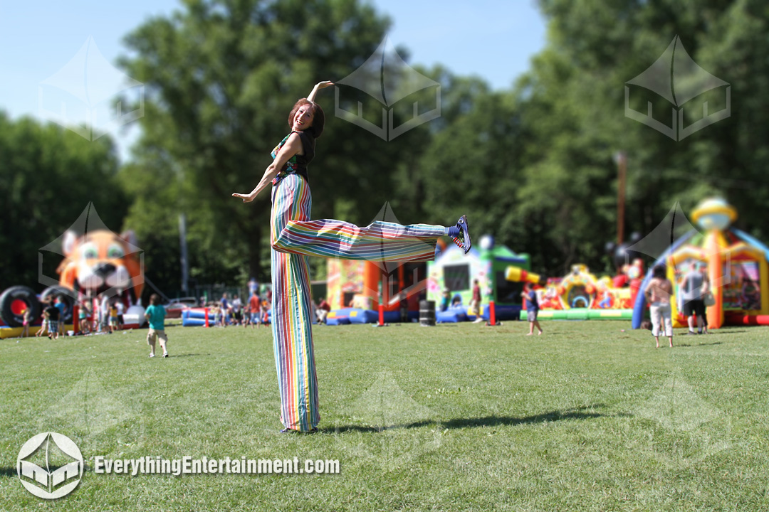 A stilt walker kicks up her feet with colorful bouncers in the background.