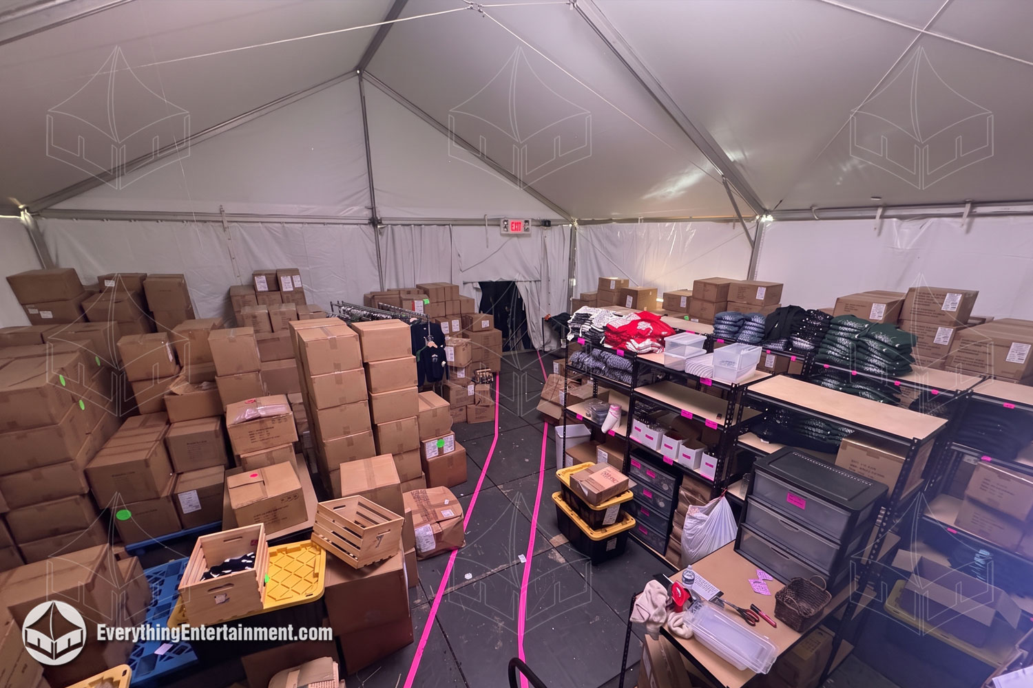 Interior of a temporary warehouse tent in active warehouse use: vinyl sidewalls, raised cassette flooring, illuminated EXIT sign, overhead string lighting, and cardboard boxes stacked beside heavy-duty industrial shelving stocked with boxed inventory under a peaked frame-tent ceiling in New Jersey